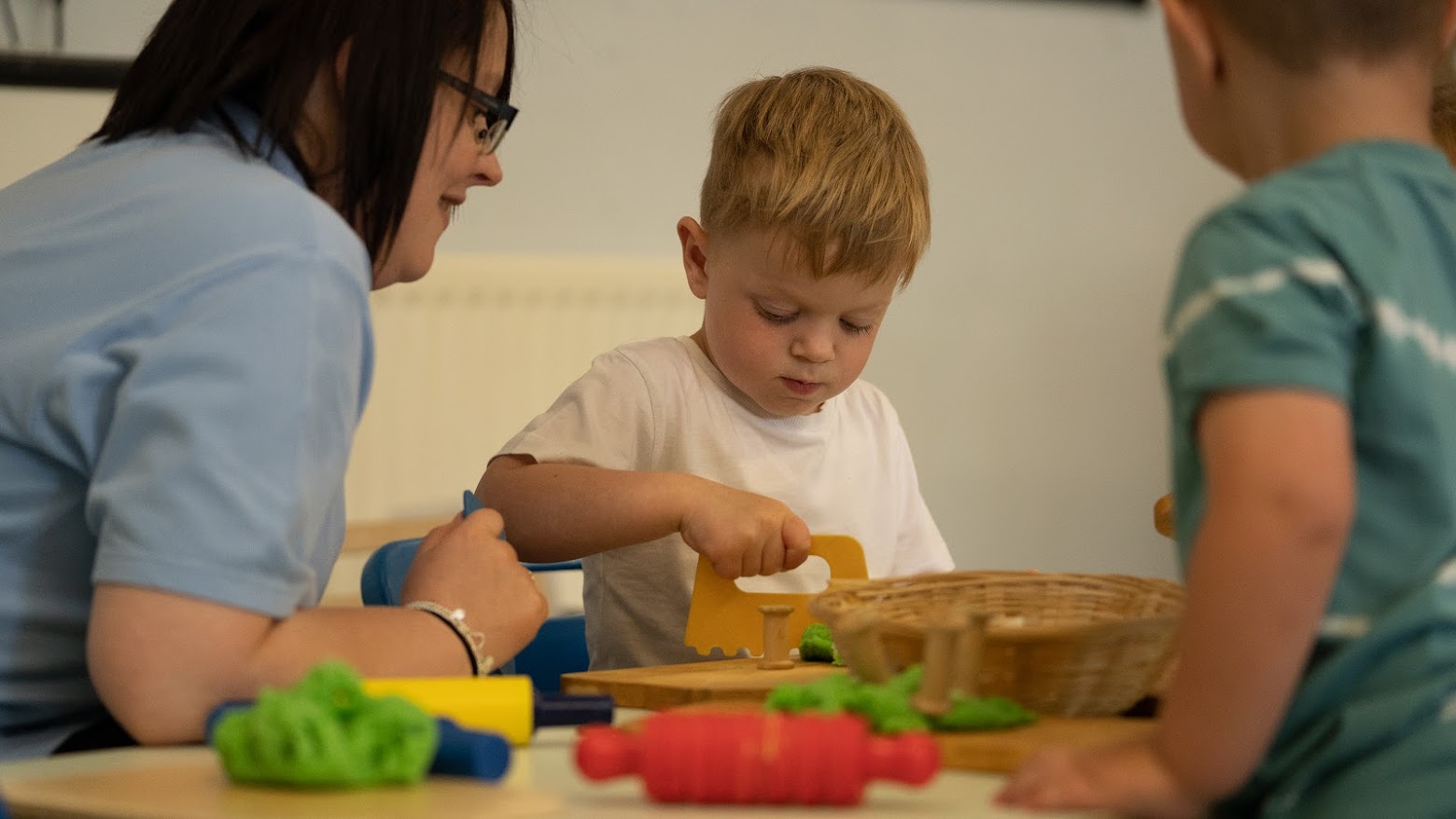 A preschool pupil is pictured playng with toys with a teacher sat next to him supervising.