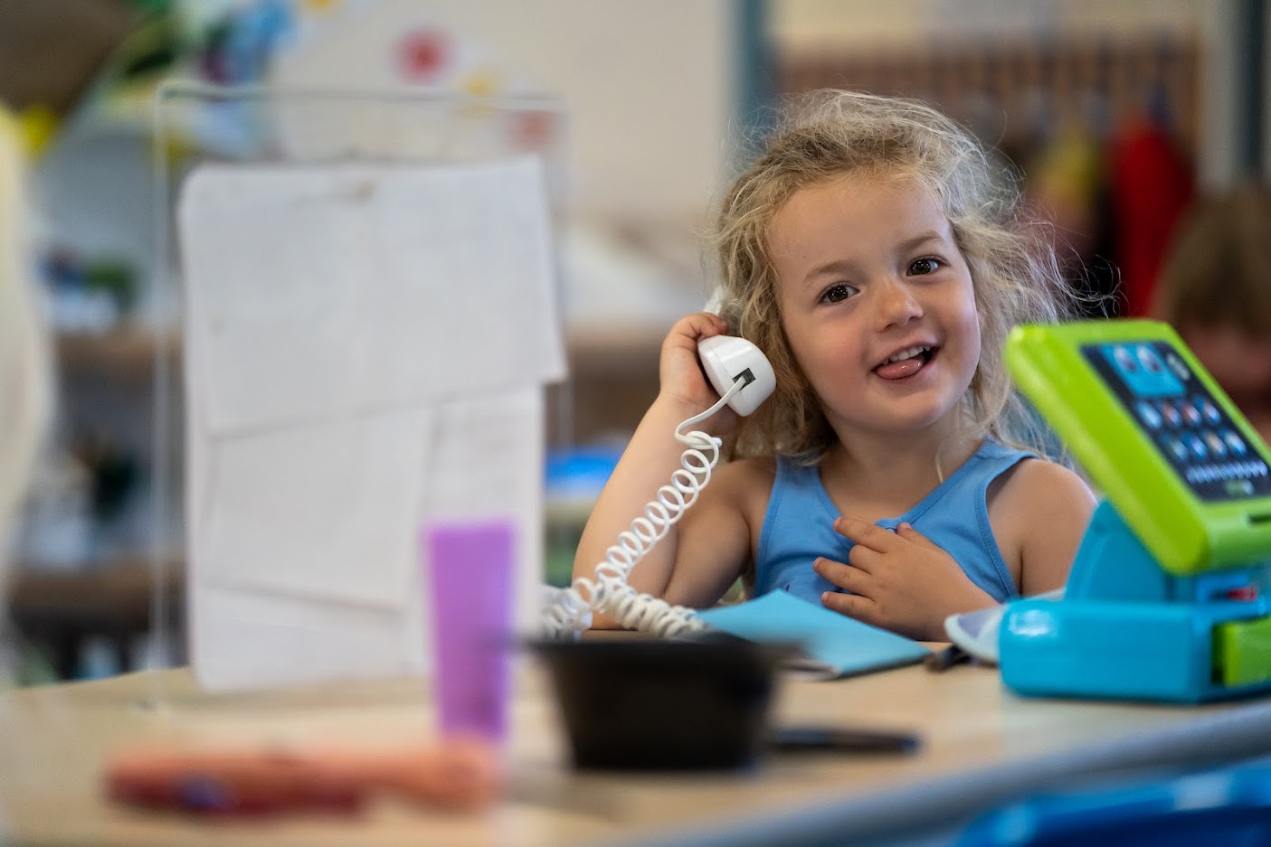 A preschool girl plays with a toy telephone to her ear and a smile on her face.