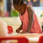 A female Nursery pupil is pictured drawing on paper with crayons whilst standing at a desk.